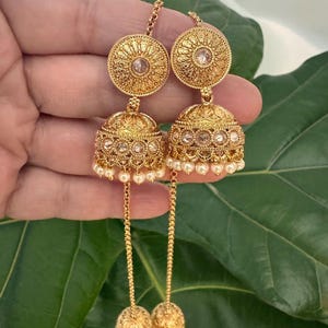 May include: Gold-toned, ornate earrings with a circular top and a bell-shaped bottom. The earrings feature intricate detailing, clear gemstones, and small white pearls. The design includes a long chain with a decorative ball at the end.