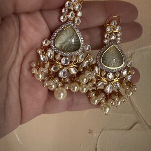 May include: A pair of gold earrings with white pearl accents. The earrings feature a teardrop-shaped design with a clear stone in the center and intricate detailing around the stone.