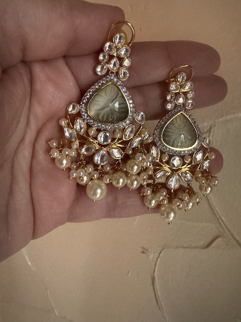 May include: A pair of gold earrings with white pearl accents. The earrings feature a teardrop-shaped design with a clear stone in the center and intricate detailing around the stone.