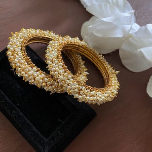 Puede incluir: Dos brazaletes de oro con cuentas de perlas blancas. Los brazaletes están apilados uno encima del otro y están sobre una caja de terciopelo negro.