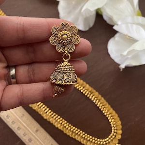 May include: Gold-toned jewelry set featuring a floral earring and a matching necklace. The earring has a flower-shaped top and a bell-shaped bottom. The necklace has a series of small, round, coin-like elements. The set is displayed against a backdrop of white flowers.