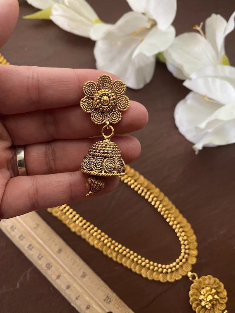 May include: Gold-toned jewelry set featuring a floral earring and a matching necklace. The earring has a flower-shaped top and a bell-shaped bottom. The necklace has a series of small, round, coin-like elements. The set is displayed against a backdrop of white flowers.