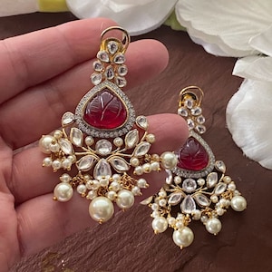 May include: A pair of gold earrings with red gemstone drops, surrounded by clear stones and pearl accents.