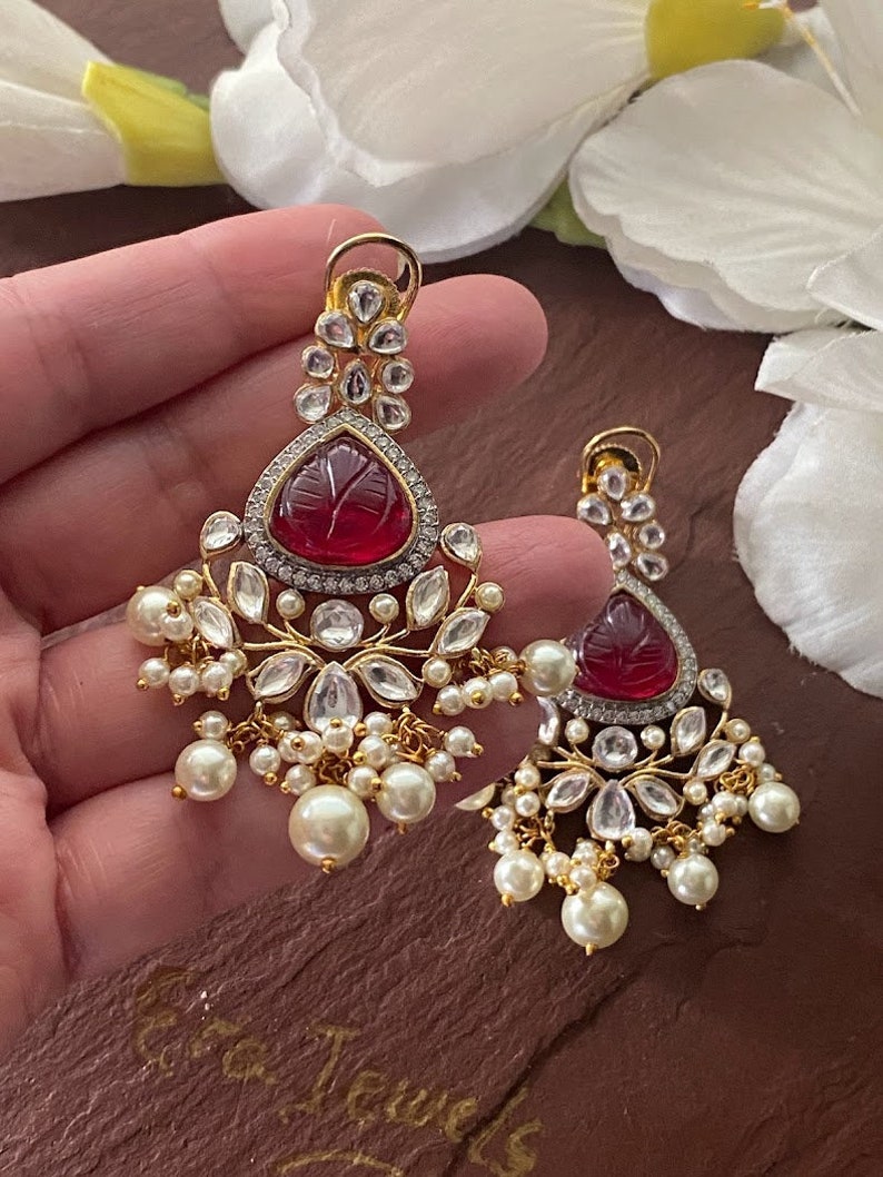 May include: A pair of gold earrings with red gemstone drops, surrounded by clear stones and pearl accents.