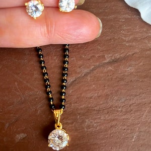 May include: A gold chain necklace with a black bead chain and a pendant featuring a large, clear, round gemstone. The necklace is paired with matching stud earrings with clear round gemstones.