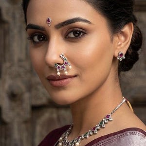 May include: A silver nose ring featuring a crescent shape with inlaid red and green stones and small dangling white pearls. The jewelry set includes matching stud earrings and a necklace with similar stone and pearl accents, worn with a bindi.