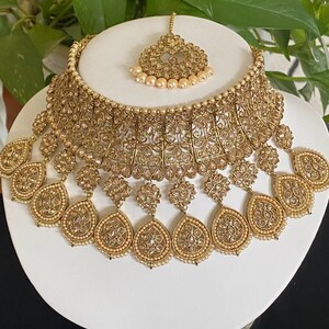 May include: A gold-tone Indian bridal necklace set with a choker style necklace, multiple teardrop-shaped pendants, and a matching maang tikka.