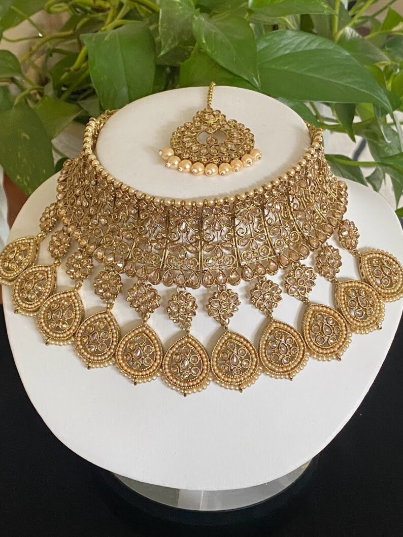 May include: A gold-tone Indian bridal necklace set with a choker style necklace, multiple teardrop-shaped pendants, and a matching maang tikka.