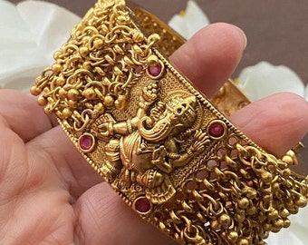 Puede incluir: Pulsera de oro con un diseño detallado de una deidad hindú, con detalles de piedras preciosas rojas y una cadena colgante con cuentas.