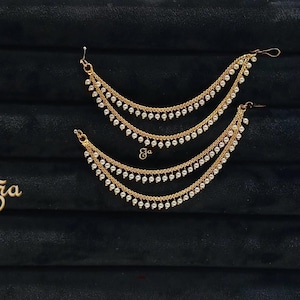May include: Gold-toned ear cuffs with multiple delicate chains and small white pearls. The ear cuffs are displayed on a black surface. A small gold-toned logo is also present.