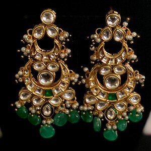 May include: A pair of gold-toned chandelier earrings with clear, round, and emerald-green accents. The earrings feature a crescent moon design with dangling emerald-green teardrop-shaped beads and small white pearls.