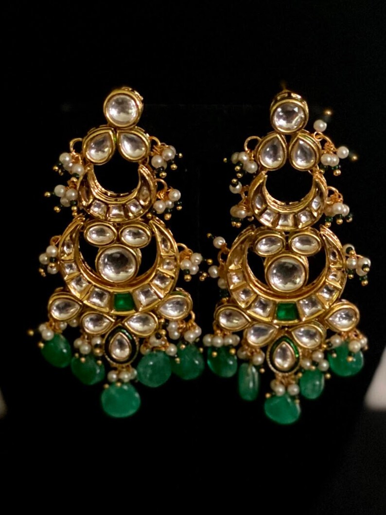 May include: A pair of gold-toned chandelier earrings with clear, round, and emerald-green accents. The earrings feature a crescent moon design with dangling emerald-green teardrop-shaped beads and small white pearls.