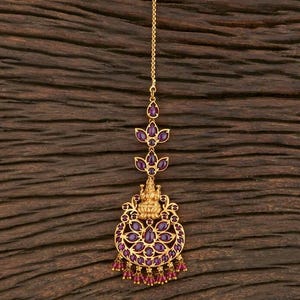 May include: A gold-toned headpiece with a chain, adorned with purple gemstones and a central design featuring a deity figure. The piece has a circular base with dangling red beads.