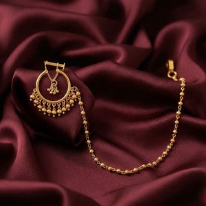 Op de afbeelding: Een goudkleurige neusring met een cirkelvormige hoepel, een bungelende centrale bedel en een reeks kleine bolvormige kralen langs de onderrand. Een lange, fijne ketting van kleine gouden kraaltjes loopt van de ring naar een sluiting.