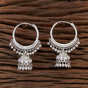 May include: A pair of silver-toned hoop earrings with intricate detailing. Each earring features a large hoop with a decorative lower edge and a bell-shaped charm with small, dangling beads. The earrings are displayed on a dark wood surface.