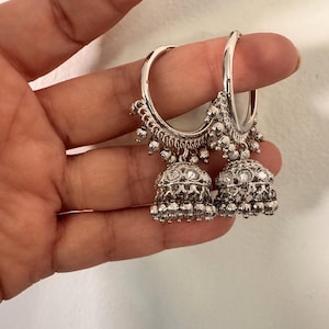 May include: A pair of silver-toned hoop earrings with intricate detailing. Each earring features a bell-shaped charm with ornate patterns and dangling beads. The earrings are held up by a finger.