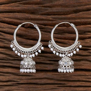 May include: A pair of silver-toned hoop earrings. Each earring features a large hoop with a decorative, scalloped edge and small white beads. A detailed, bell-shaped charm with more white beads hangs from each hoop.
