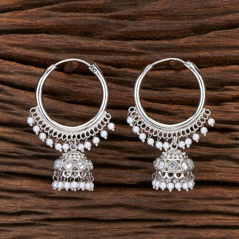 May include: A pair of silver-toned hoop earrings. Each earring features a large hoop with a decorative, scalloped edge and small white beads. A detailed, bell-shaped charm with more white beads hangs from each hoop.