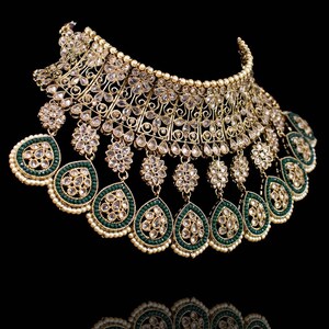 May include: A gold and green statement necklace with a large, ornate design. The necklace features a central piece with a teardrop shape and multiple smaller teardrops hanging from it. The necklace is decorated with clear stones and green beads.