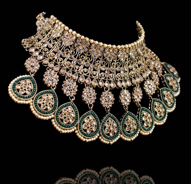 May include: A gold and green statement necklace with a large, ornate design. The necklace features a central piece with a teardrop shape and multiple smaller teardrops hanging from it. The necklace is decorated with clear stones and green beads.
