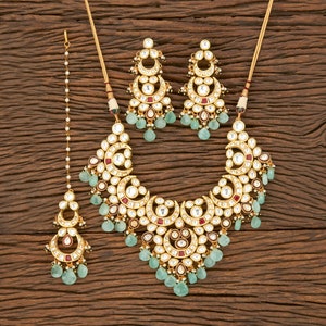 May include: A gold-toned jewelry set featuring a necklace, earrings, and a headpiece. The necklace and earrings have a crescent moon design with white and red accents, and teardrop-shaped green gems. The headpiece has a similar design.