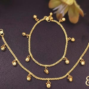 Puede incluir: Juego de tobillera y pulsera doradas. La joyer&iacute;a presenta un delicado dise&ntilde;o de cadena con peque&ntilde;os dijes de campana colgantes. Las piezas se exhiben sobre una superficie oscura, con un acento floral en el fondo. La tobillera y la pulsera probablemente sean de metal dorado.