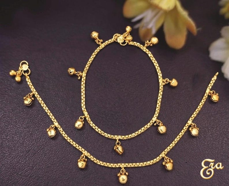 Puede incluir: Juego de tobillera y pulsera doradas. La joyer&iacute;a presenta un delicado dise&ntilde;o de cadena con peque&ntilde;os dijes de campana colgantes. Las piezas se exhiben sobre una superficie oscura, con un acento floral en el fondo. La tobillera y la pulsera probablemente sean de metal dorado.