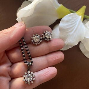 May include: A silver necklace with a black beaded chain and a pendant featuring a large round clear stone surrounded by smaller stones. Matching earrings with a similar design are also shown.