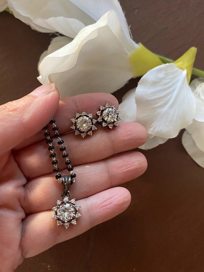 May include: A silver necklace with a black beaded chain and a pendant featuring a large round clear stone surrounded by smaller stones. Matching earrings with a similar design are also shown.