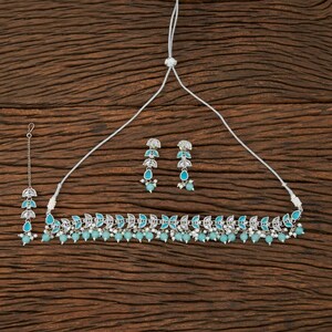 May include: A silver-toned jewelry set featuring a necklace, earrings, and a headpiece. The necklace and headpiece have a leaf-like design with turquoise-colored accents and small white beads. The earrings have a similar design with turquoise teardrop-shaped gems.