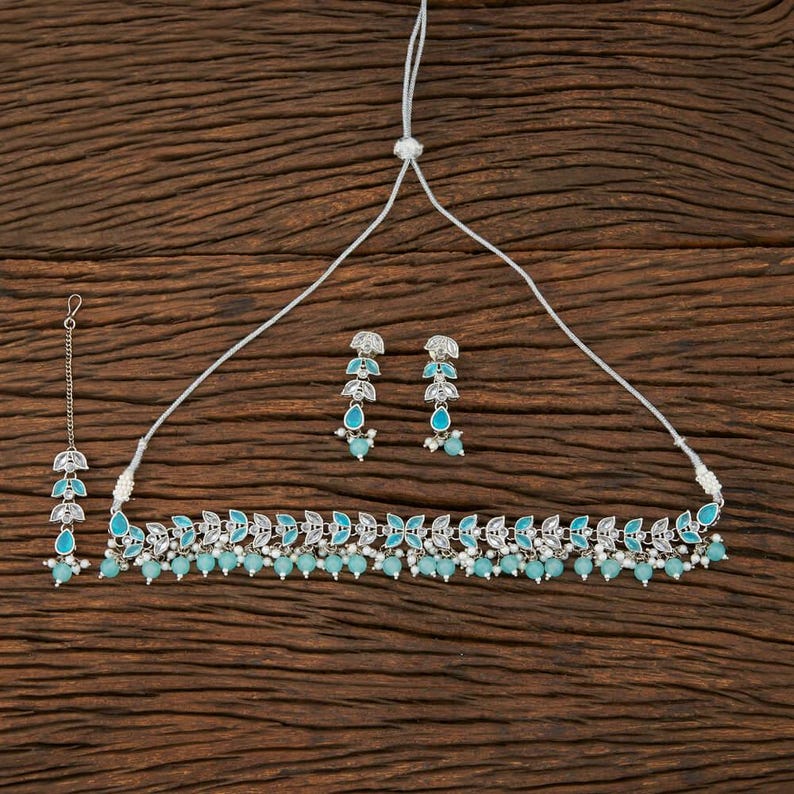 May include: A silver-toned jewelry set featuring a necklace, earrings, and a headpiece. The necklace and headpiece have a leaf-like design with turquoise-colored accents and small white beads. The earrings have a similar design with turquoise teardrop-shaped gems.