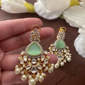 May include: A pair of gold earrings with a green stone and pearl accents. The earrings have a floral design and are dangling.