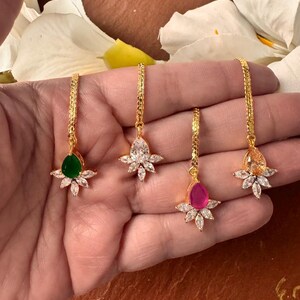 Puede incluir: Cuatro pendientes colgantes dorados con piedras preciosas de diferentes colores. Cada pendiente tiene una cadena y un diseño floral con una piedra central verde, transparente, rosa y naranja. Los pendientes se sostienen en una mano.