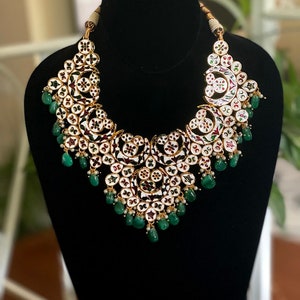 May include: An ornate necklace with a gold-colored setting, featuring white enamel and intricate floral designs. The necklace is adorned with small, round red, green, and white accents, and teardrop-shaped green gemstones. The necklace is displayed on a black mannequin.
