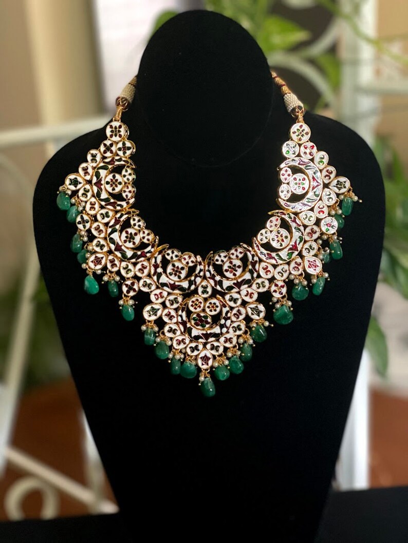 May include: An ornate necklace with a gold-colored setting, featuring white enamel and intricate floral designs. The necklace is adorned with small, round red, green, and white accents, and teardrop-shaped green gemstones. The necklace is displayed on a black mannequin.