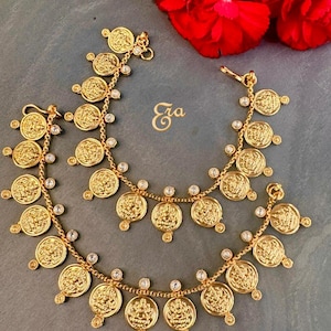 May include: Gold-toned anklets featuring coin-shaped charms with intricate designs and small, clear gemstones. The anklets are displayed on a gray surface with red flowers in the background. The word "Era" is also visible.