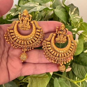 May include: A pair of gold-tone earrings with a crescent moon design and a small, detailed figure of a deity at the top. The earrings have a dangling ball at the bottom.