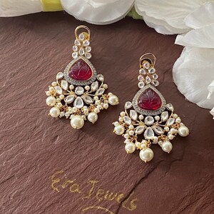 May include: A pair of gold earrings with red and white stones. The earrings have a teardrop shape and are decorated with pearl accents. The earrings are on a brown surface with the text "Era Jewels" written in gold.