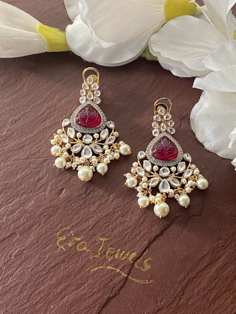 May include: A pair of gold earrings with red and white stones. The earrings have a teardrop shape and are decorated with pearl accents. The earrings are on a brown surface with the text "Era Jewels" written in gold.