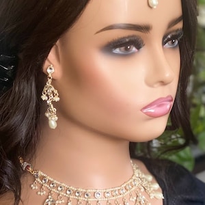 May include: A gold and white bridal jewellery set with a choker necklace, earrings, and a tikka. The necklace features a double row of pearls and a dangling design with pearl accents. The earrings are long and feature a similar design to the necklace. The tikka is a traditional Indian headpiece with a pearl drop.