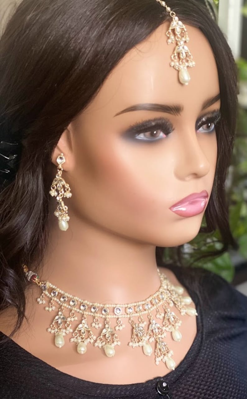 May include: A gold and white bridal jewellery set with a choker necklace, earrings, and a tikka. The necklace features a double row of pearls and a dangling design with pearl accents. The earrings are long and feature a similar design to the necklace. The tikka is a traditional Indian headpiece with a pearl drop.