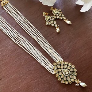 May include: A white pearl necklace with a gold pendant featuring a green and gold design with clear stones. The necklace is layered with multiple strands of pearls. Matching earrings with a similar design are also included.