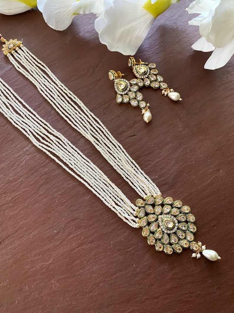 May include: A white pearl necklace with a gold pendant featuring a green and gold design with clear stones. The necklace is layered with multiple strands of pearls. Matching earrings with a similar design are also included.