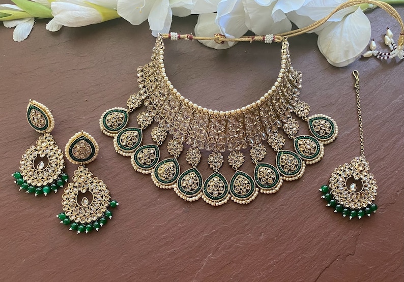 May include: A gold and green Indian bridal necklace set with a large, ornate choker, matching earrings, and a maang tikka. The necklace features intricate floral designs and is adorned with sparkling stones and pearls.