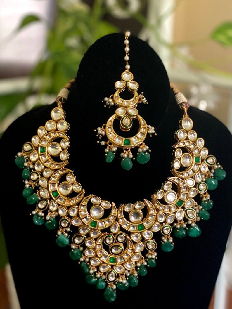 May include: A gold-toned necklace and headpiece set. The necklace features a crescent moon design with clear and green accents, and dangling green beads. The headpiece has a similar design with dangling pearls.