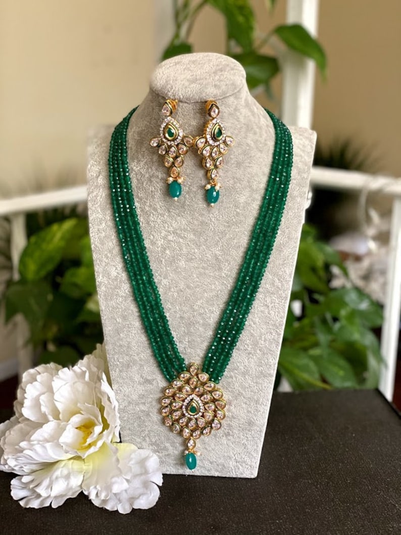 May include: A green beaded necklace with a gold pendant and matching earrings. The pendant features a circular design with white stones and a green center stone. The earrings are also circular with white stones and a green center stone.