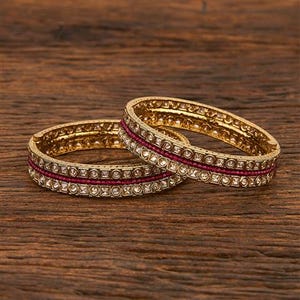 May include: Two gold-colored bangles with a central band of small, red gemstones and a border of clear, square-cut stones. The bangles are set against a dark wood grain background.