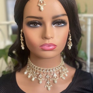 May include: A gold and white bridal jewellery set with a choker necklace, earrings, and a tikka. The jewellery is adorned with pearls and rhinestones.
