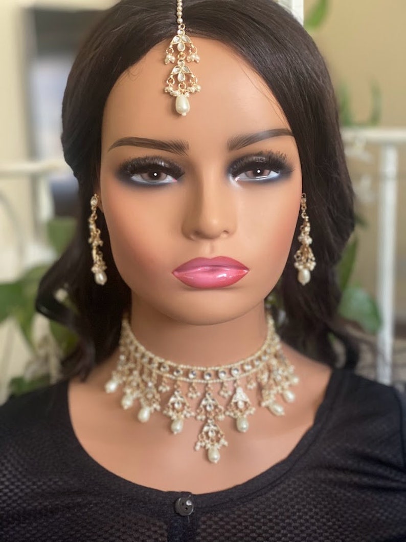 May include: A gold and white bridal jewellery set with a choker necklace, earrings, and a tikka. The jewellery is adorned with pearls and rhinestones.