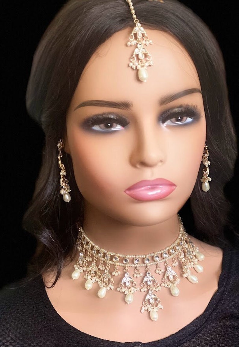 May include: A white and gold bridal jewellery set with pearl accents. The set includes a choker necklace, earrings, and a tikka.
